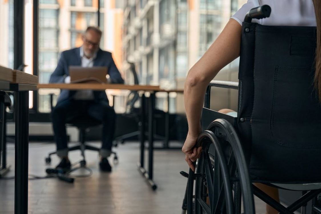 Two people working in an office, one at a desk and the other seated in a wheelchair.