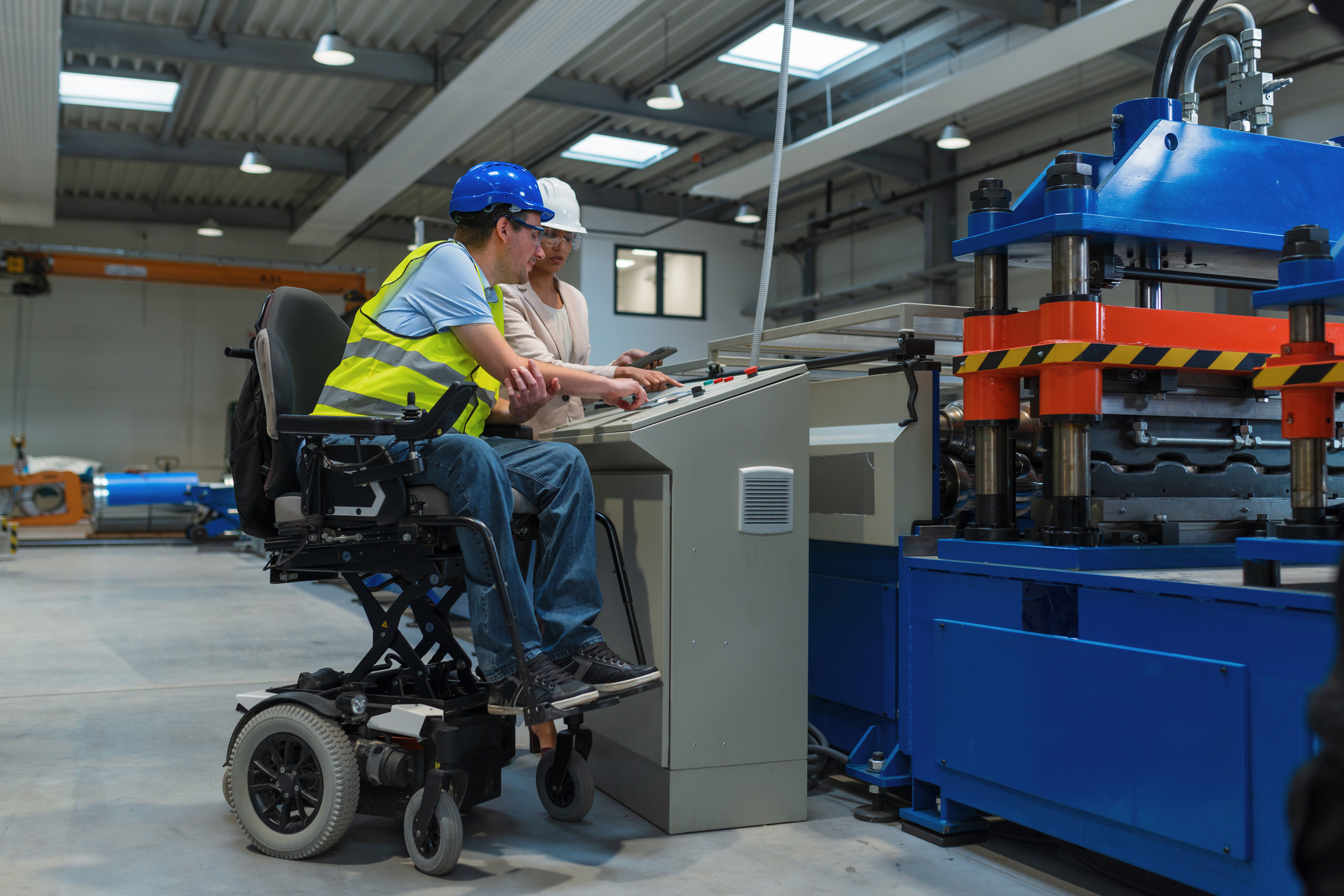 Man in elevating wheelchair works with a female co-worker at a factory control panel.